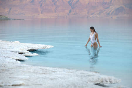 Middle eastern female model in a white one-piece swimsuit stands waist-deep in vibrant blue mineral-rich water at the famous Dead Sea