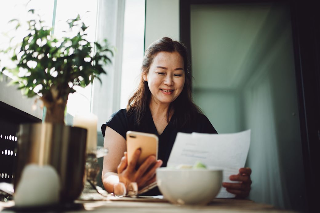A middle-aged Asian woman looking at her phone and a bank statement while sitting at her kitchen table.