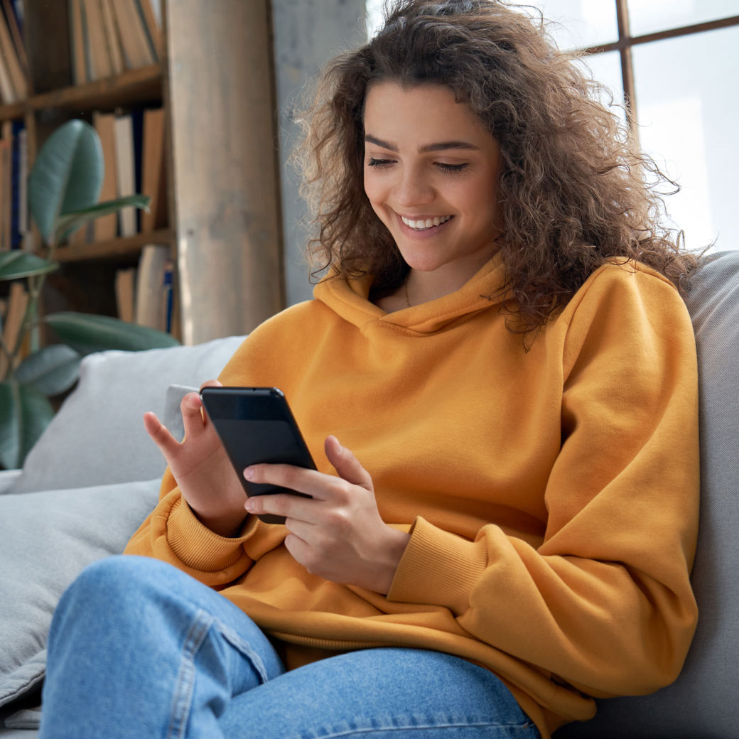 A girl in an orange sweatshirt sitting on the couch at home, smiling while looking at her phone.