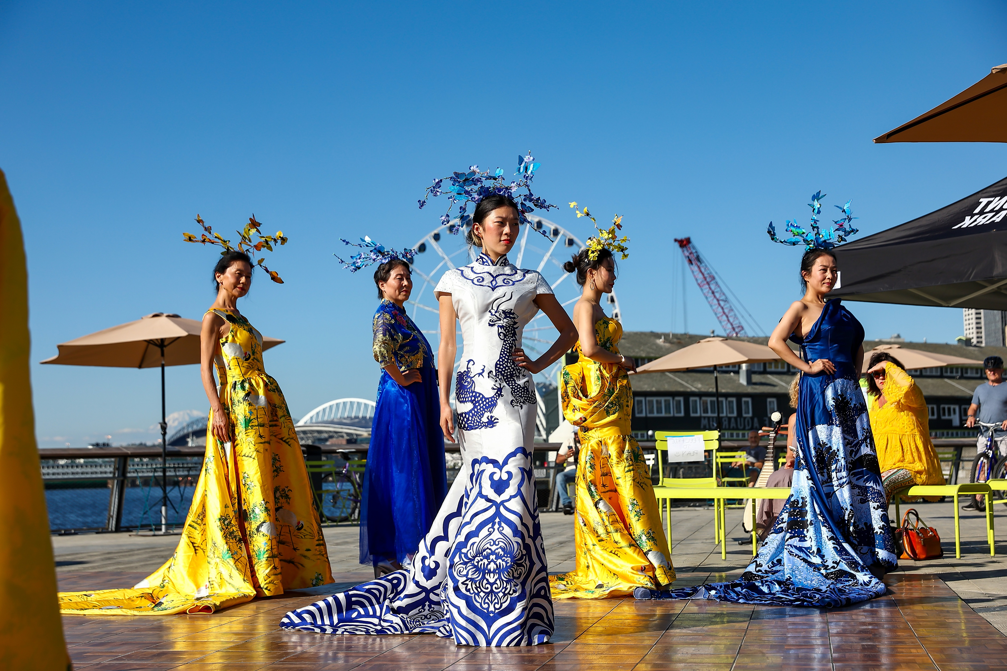 Five figures stand outdoors on a sunny day, wearing elaborate, colorful dresses with long trains in blue, yellow, and white. Each model wears an ornate headpiece. The Seattle Waterfront is visible in the background."