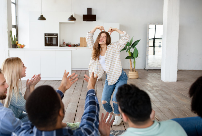 A group of people playing Charades in a living room, with a wide plank wooden floor, white walls and black light fixtures