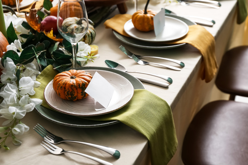 Table set for Thanksgiving with small pumpkins at each place setting, with table runners in green and orange and place cards, wine glass, flatware and dishes.