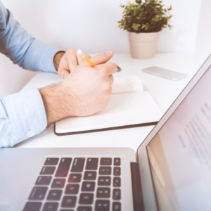 What will BT’s broadband and call price rises mean for you? Image of a man's hands holding a pen by a notebook. Laptop and plant.