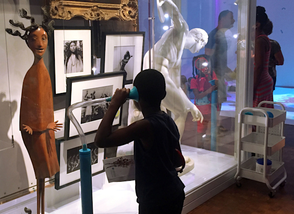 A young boy listens to artwork-related audio pieces through a flexible ear horn tube connected to a transducer, which is part of the Artquarium.