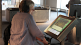 Older woman listens to audio and looks at map on Floods Navigator kiosk in gallery.