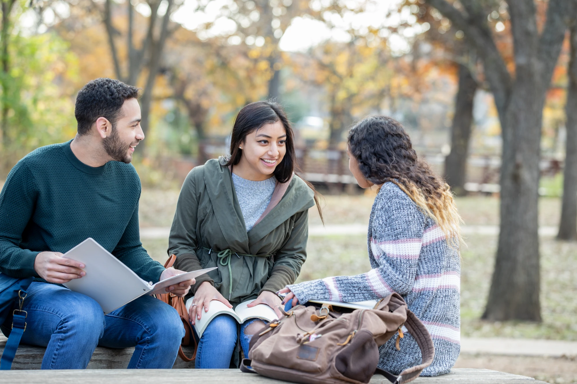 Three smiling students seated and conversing outdoors on campus in autumn