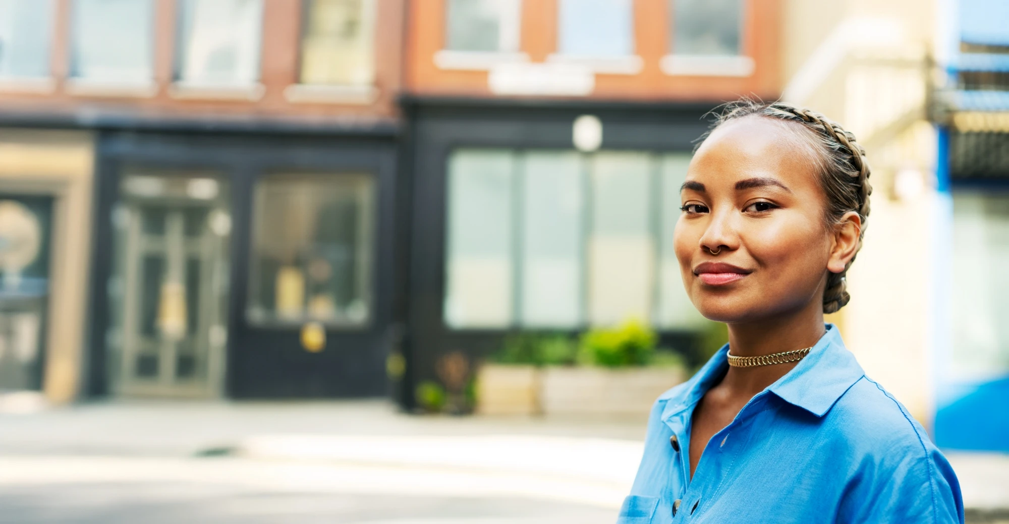 Student in blue blouse stands outdoors in front of storefronts and smiles