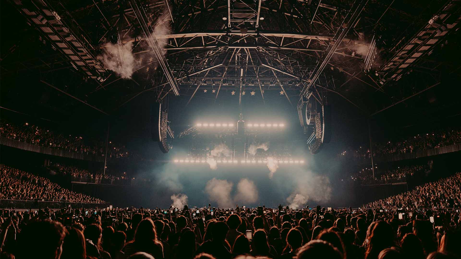The AFAS Dome, photographed from the back row. The stage lights come on and many fans on the floor have their phones raised in the air.