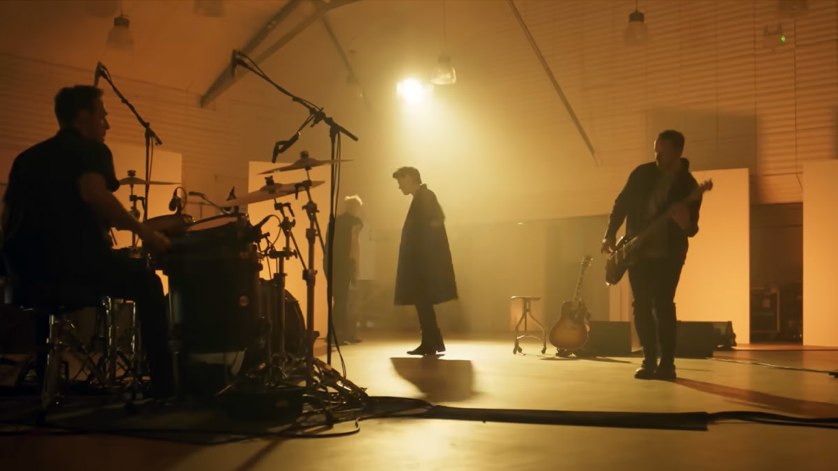 The Script repeteert in een sfeervol verlichte studio tijdens de opname van ‘Man in the Arena’. Frontman Danny O’Donoghue staat centraal in beeld, omringd door drummer en gitarist.