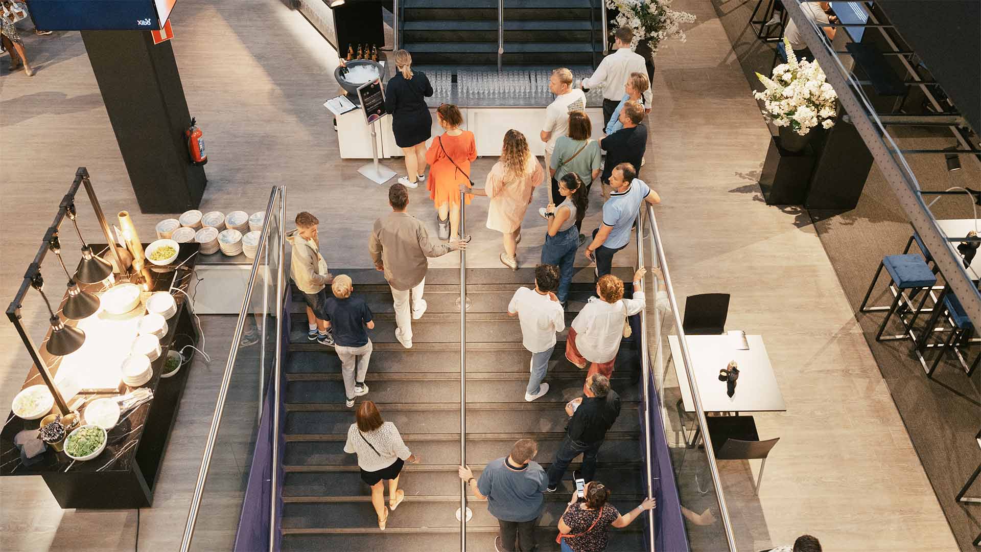 Een bovenaanzicht van mensen die de trap op en af gaan in het Hospitality Center, met een buffet en drankstation zichtbaar.