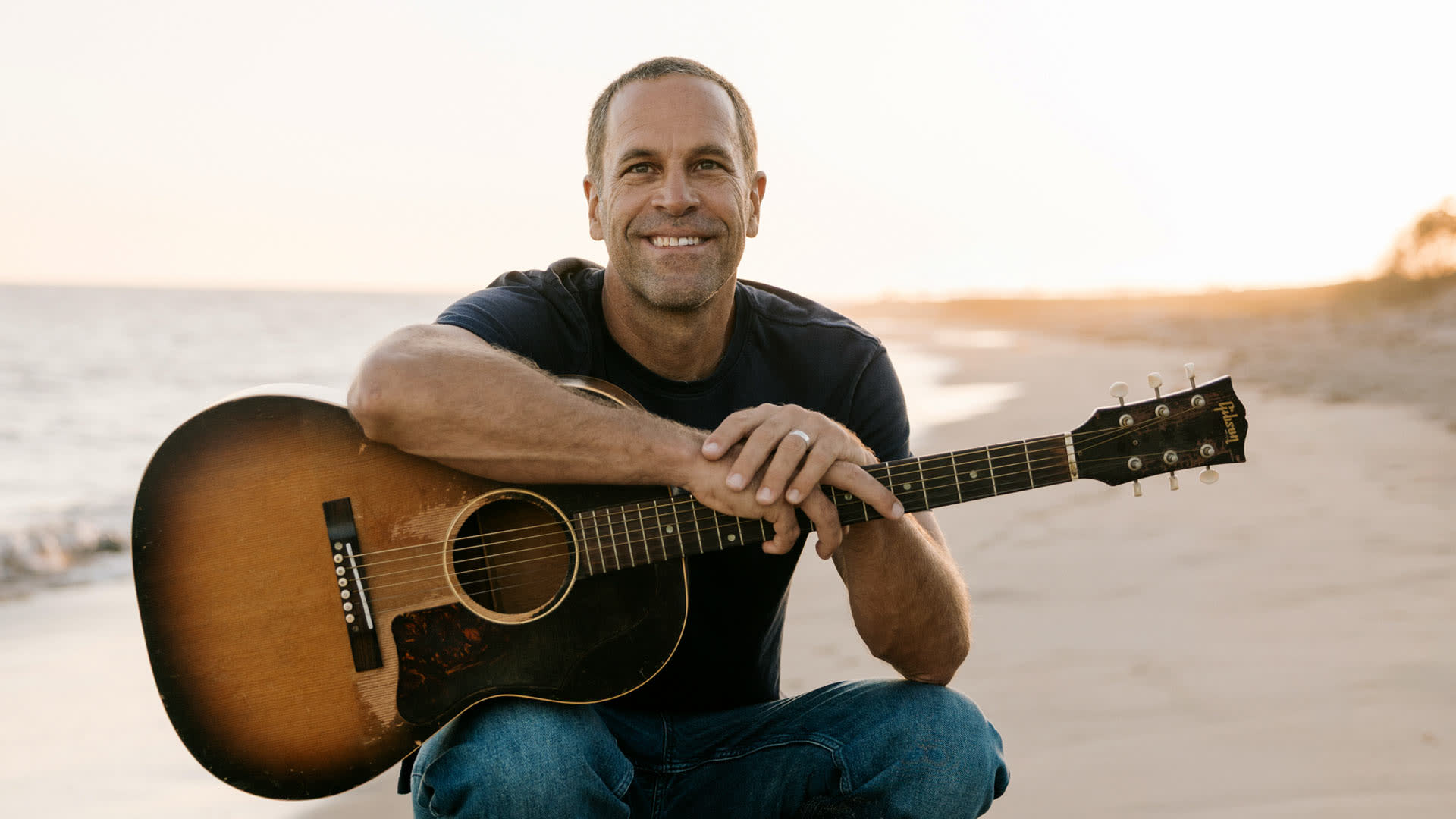 Jack Johnson met een klassieke gitaar op het strand met op de achtergrond de zee.