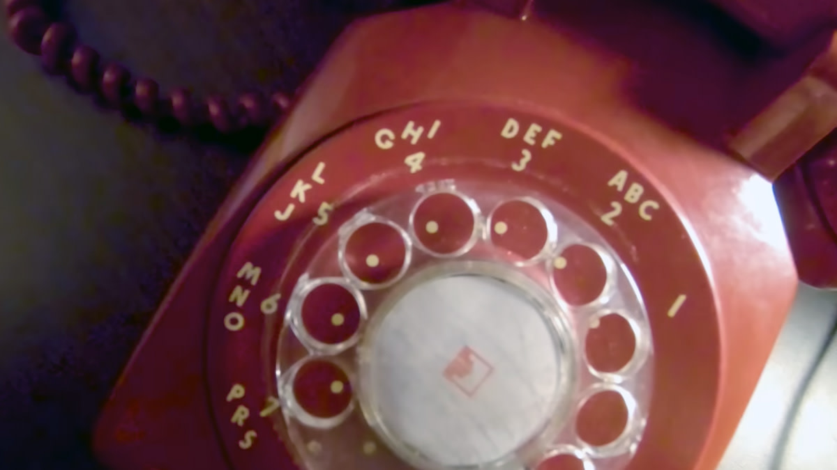 Close-up van een rode draaischijftelefoon met letters en cijfers rond de kiesschijf, badend in paars-roze licht.