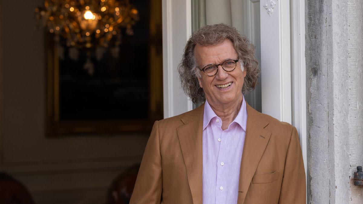 André Rieu staat in een net pak voor een open deur met binnen een prachtige klassieke luster.
