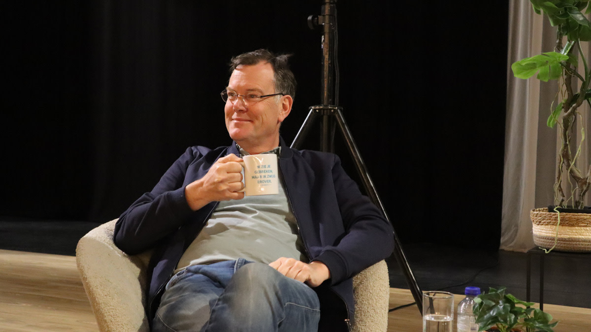 Comedian Arnout Van den Bossche zit ontspannen in een stoel met een mok in de hand, glimlachend tijdens een gesprek op het podium.