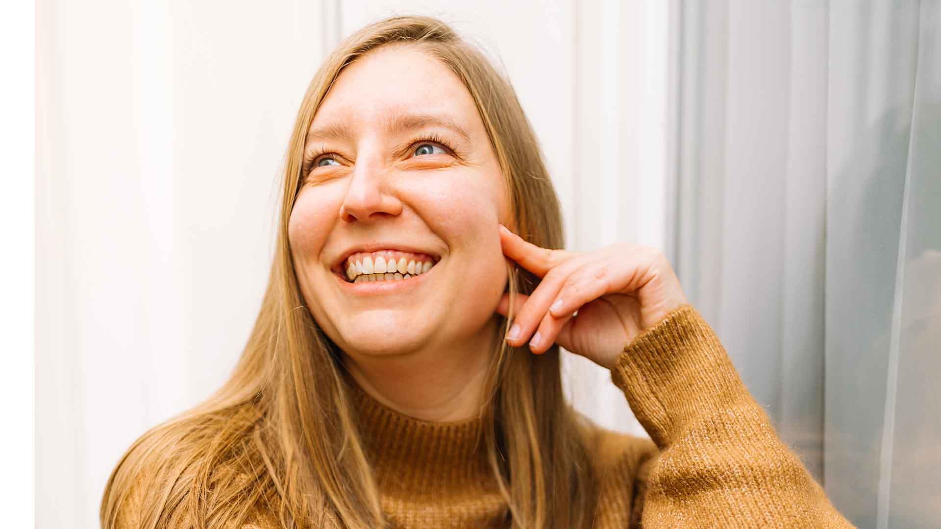 Een lachende vrouw met lang blond haar en blauwe ogen, gekleed in een bruine coltrui, kijkt omhoog en naar rechts. Ze heeft haar linkerhand bij haar wang. De achtergrond is onscherp en licht van kleur.