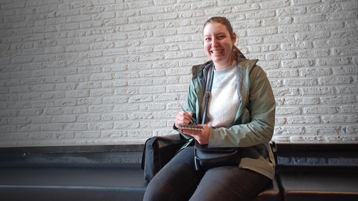 Een lachende vrouw zit op een bank voor een witte bakstenen muur en houdt een notitieboekje en pen vast.