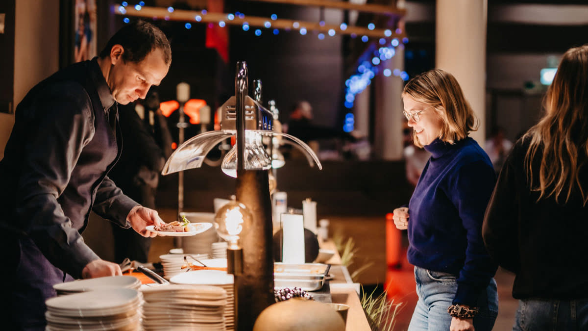 Een medewerker schept eten op aan een verzorgd buffet terwijl een vrouw glimlachend toekijkt in een sfeervol verlichte VIP-ruimte.