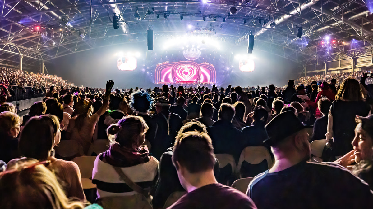 Publiek viert mee tijdens Het Schlagerfestival in een uitverkochte Trixxo Arena. Duizenden bezoekers kijken richting het kleurrijk verlichte podium, terwijl de zaal baadt in een feestelijke sfeer met lichtshows, glitter en meezingmomenten.