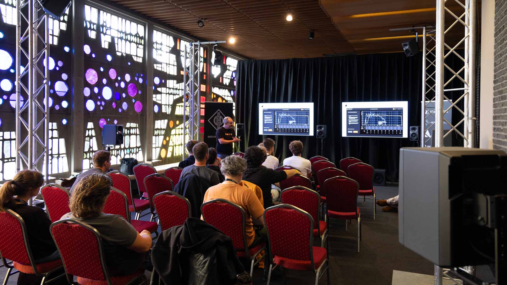 In de zaal staan verschillende rijen met rode stoelen opgesteld. Een man geeft een presentatie voor een tiental mensen.