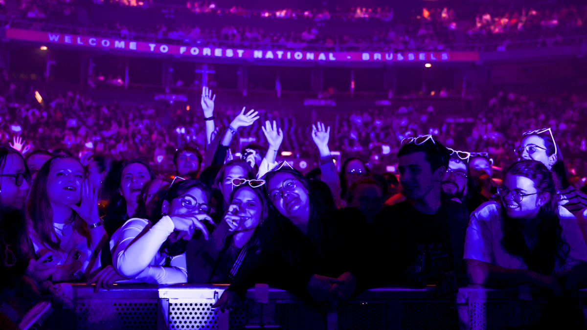 Publiek dicht bij de barricade in Vorst Nationaal tijdens het concert van Helena, verlicht in paarse en blauwe podiumkleuren. Fans lachen, zingen en steken hun handen in de lucht terwijl de volle zaal op de achtergrond zichtbaar is.