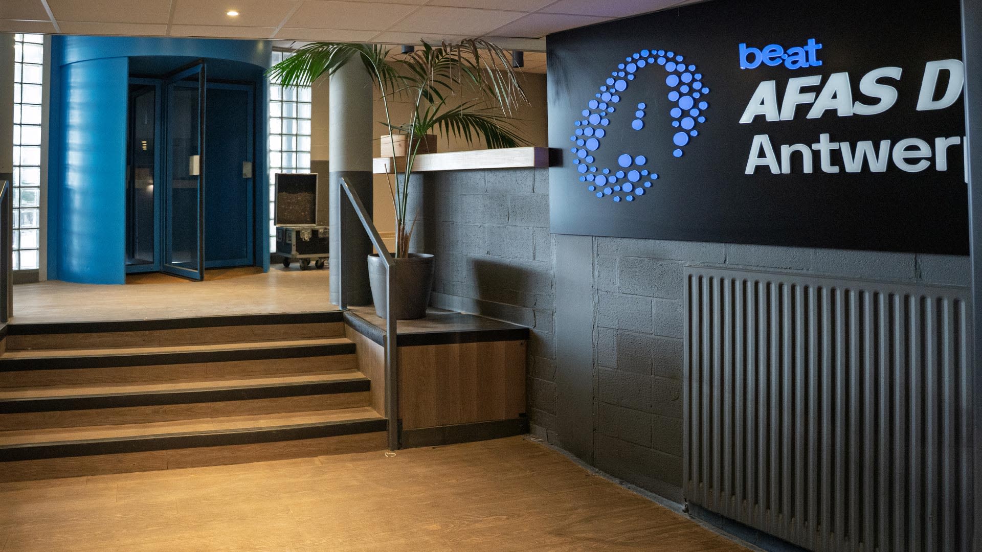 Het huidige backstageonthaal in de AFAS Dome in Antwerpen, met een blauwe inkomhal, houten trap, planten en het verlichte be•at-logo op de muur.