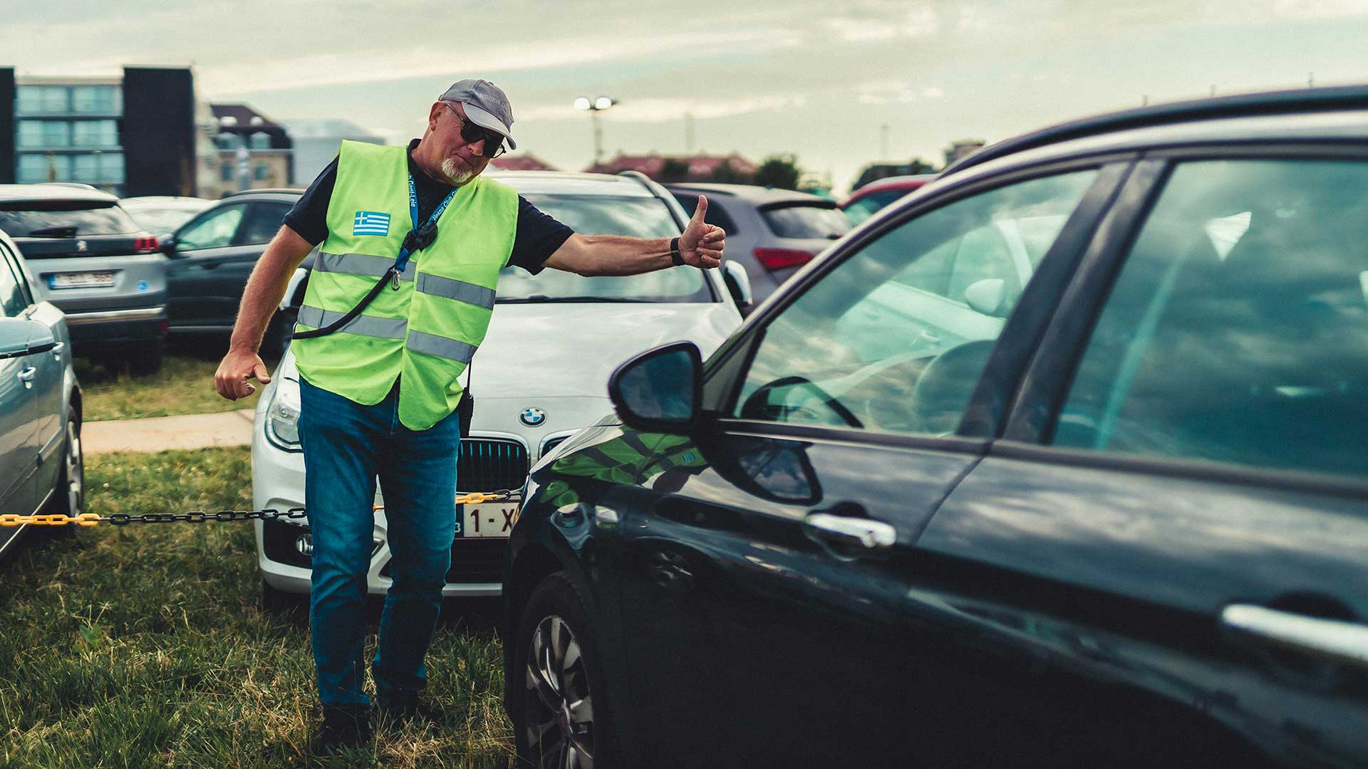 Een verkeersbegeleider in fluohesje en petje wijst met een duim omhoog naar een auto op een druk parkeerterrein. Rondom staan meerdere wagens geparkeerd op gras.