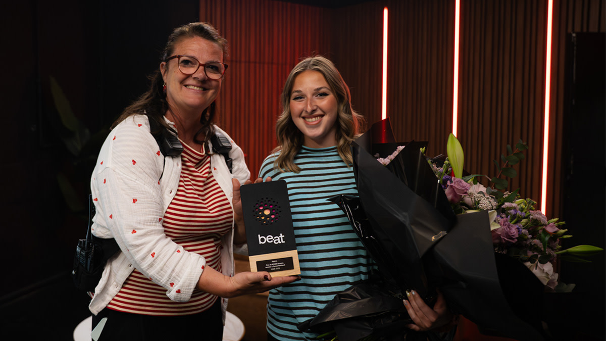 Coralie Berael overhandigt de be•at Award aan zangeres Helena, die glimlachend een boeket bloemen vasthoudt, tijdens een moment achter de schermen.
