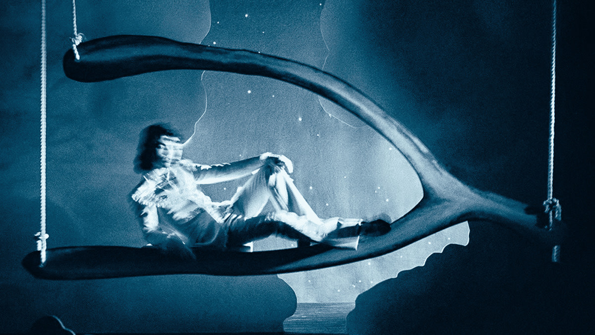 Artwork for Conan Gray's new single 'The Best': a figure rests dreamily inside a large wishbone-shaped swing, bathed in a blue, ethereal glow with scattered specks of light.