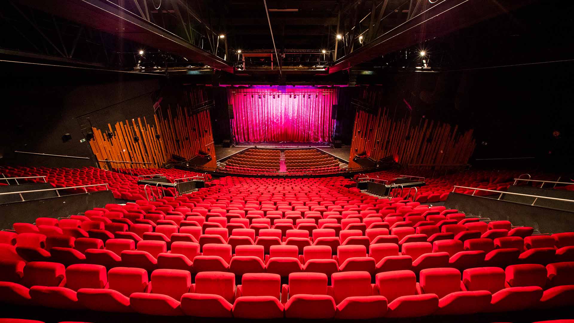Een lege zaal, bekeken van de bovenste rijen. Op het podium is een gesloten gordijn te zien en in de zaal staan duizenden lege rode stoelen.