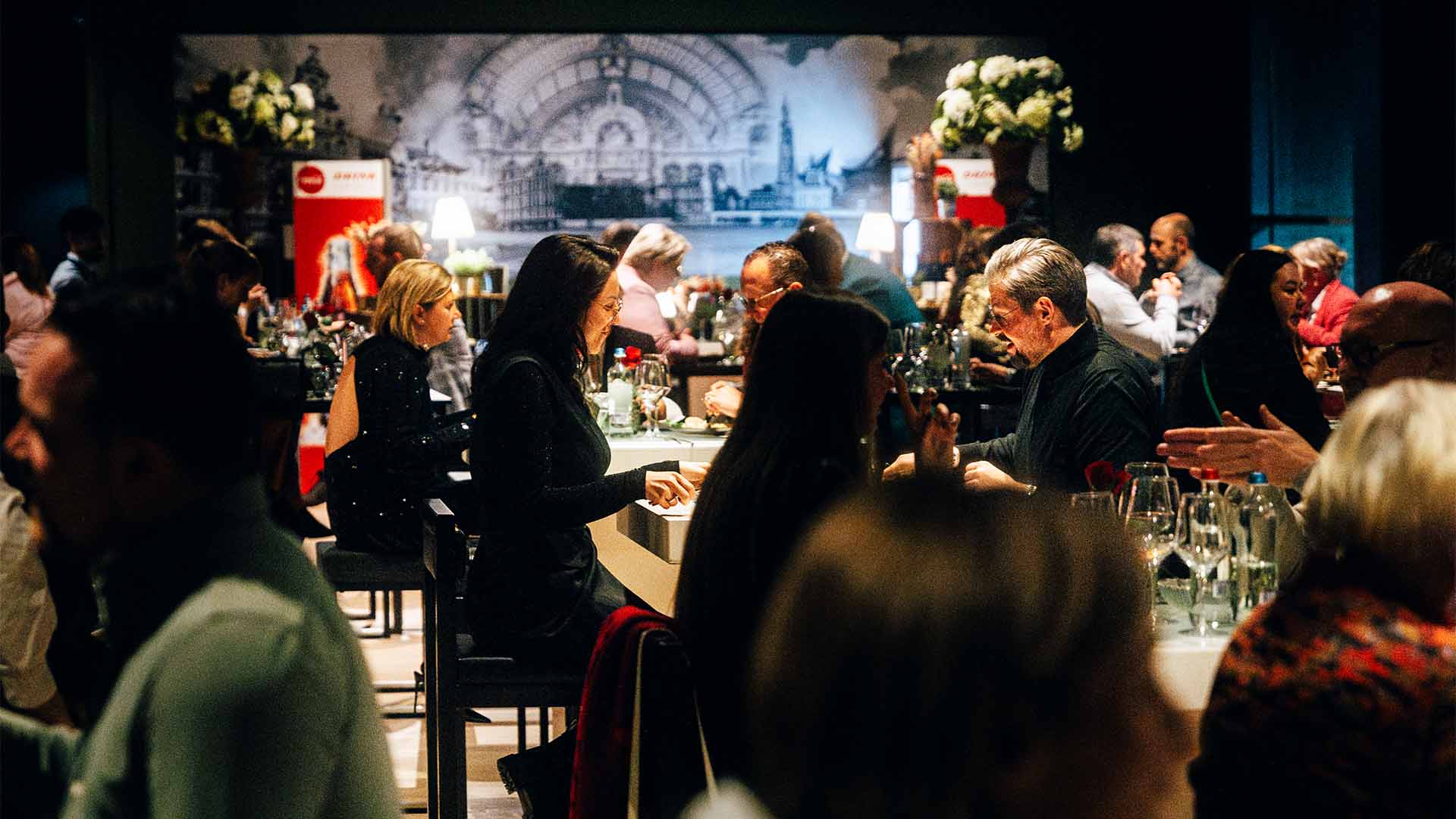 Gasten dineren en converseren aan tafels in een sfeervol verlichte evenementenruimte, met een groot wandpaneel op de achtergrond.