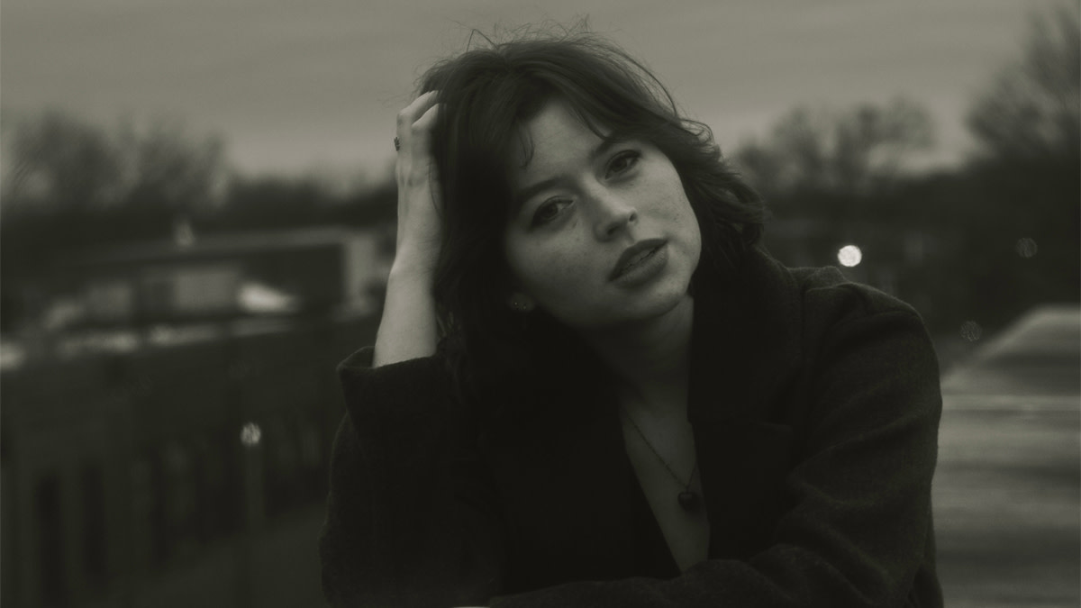 Portrait en noir et blanc de Josie Edwards, assise à l’extérieur, une main dans les cheveux, le regard tourné au loin.