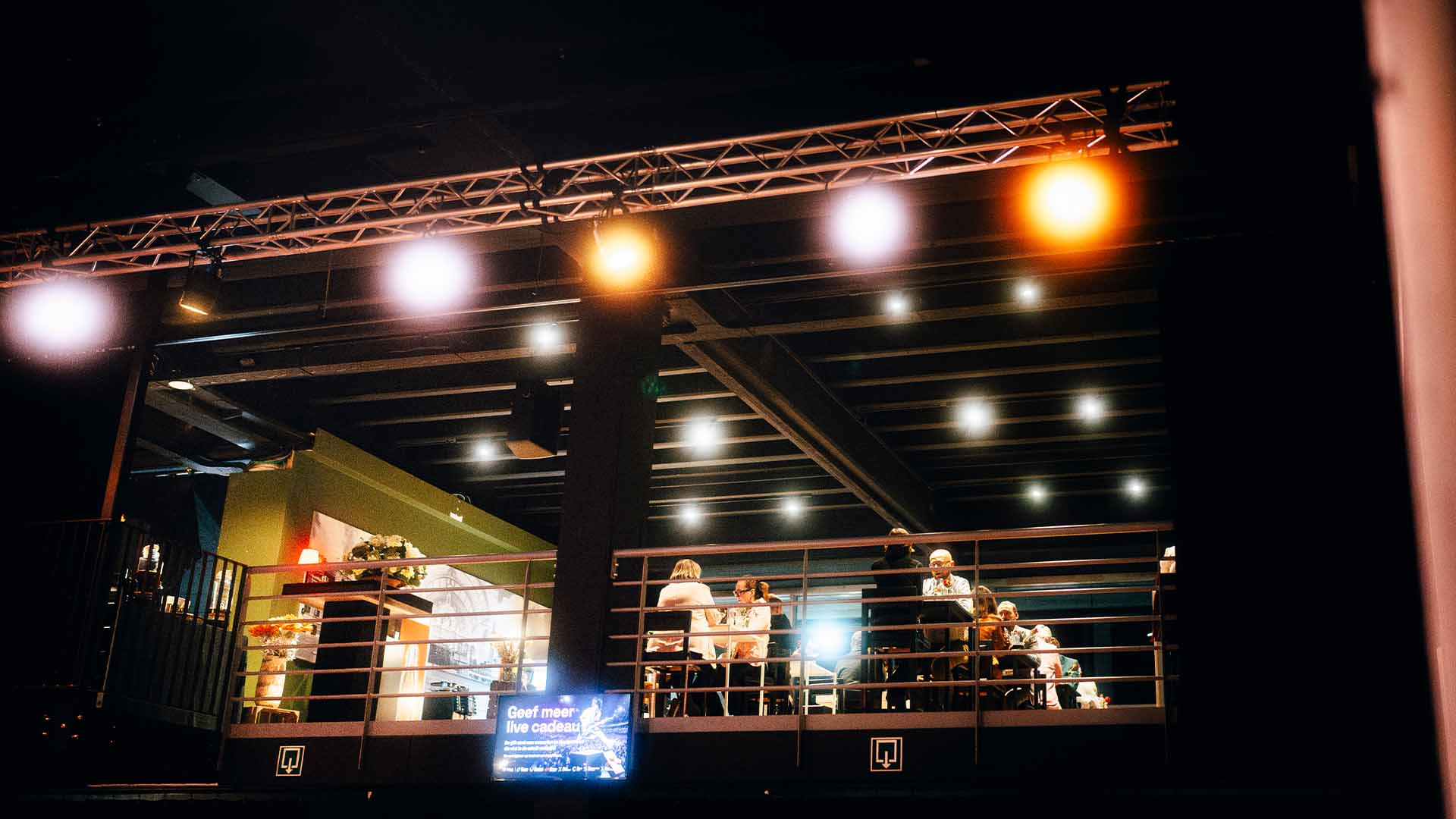 Een scène vanuit een lager perspectief die een bovenverdieping van de evenementenruimte toont met gasten aan tafels en een verlichte reclamebanner.