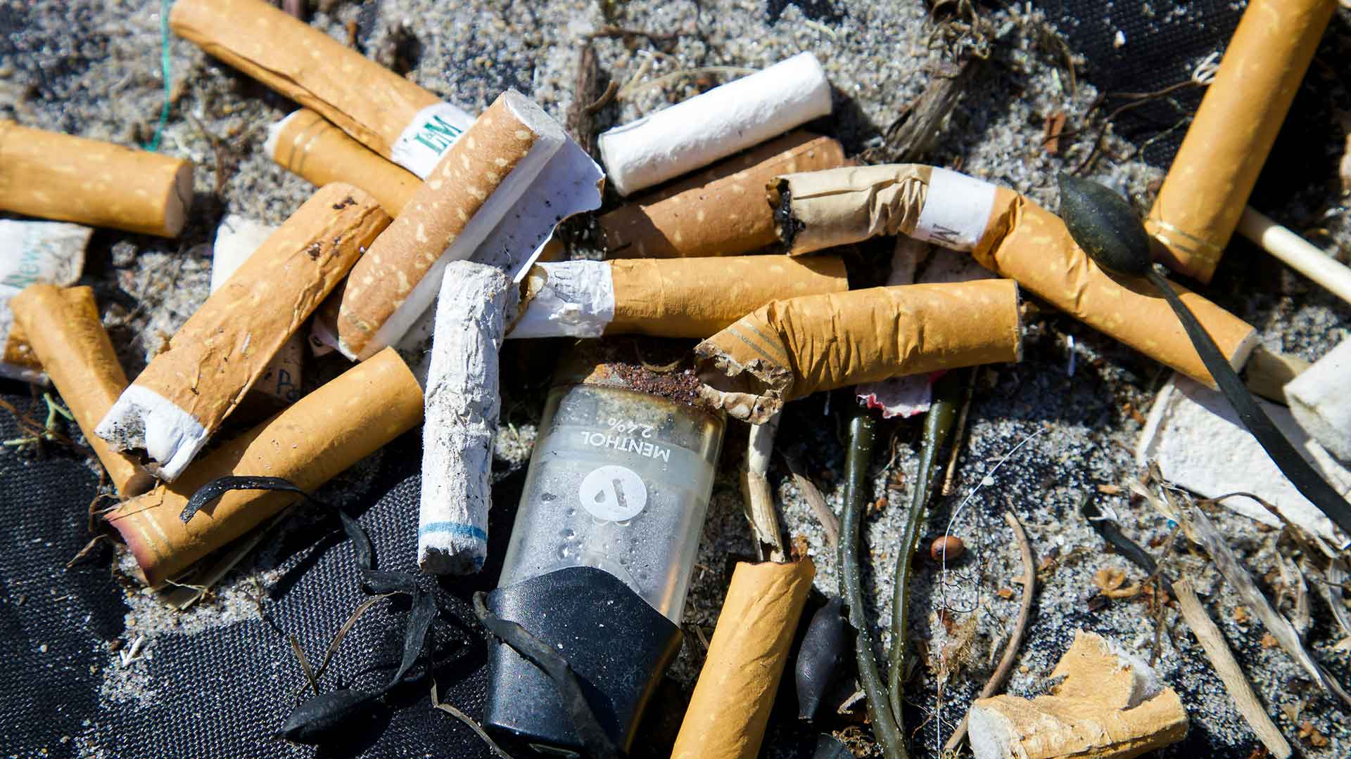 Un tas de mégots, une cigarette électronique mentholée cassée et d'autres déchets éparpillés sur le sable.