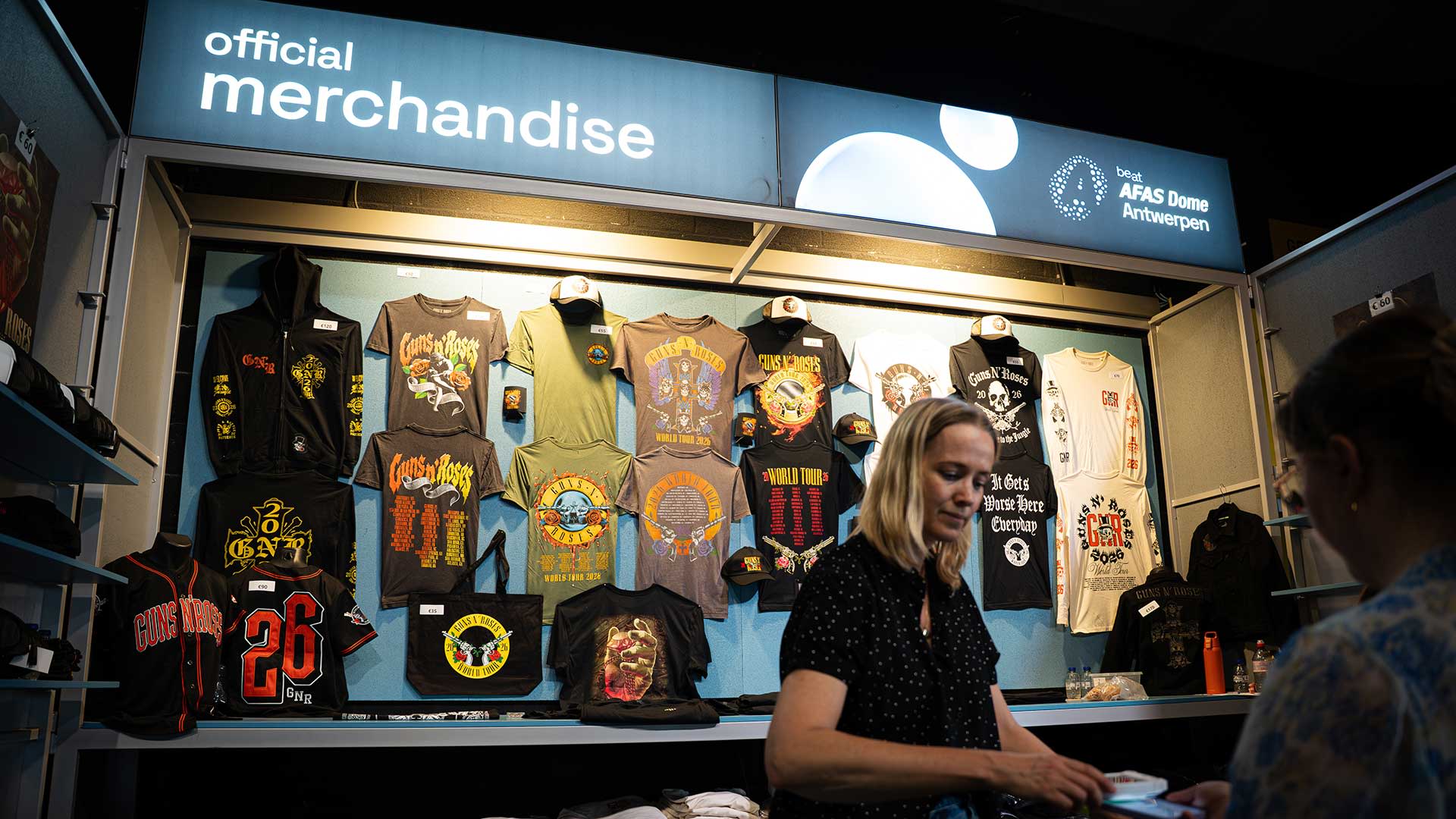Merchandise stand in the AFAS Dome with an ‘Official merchandise’ sign, a wall displaying T‑shirts and clothing, and a staff member behind the counter handling a purchase.