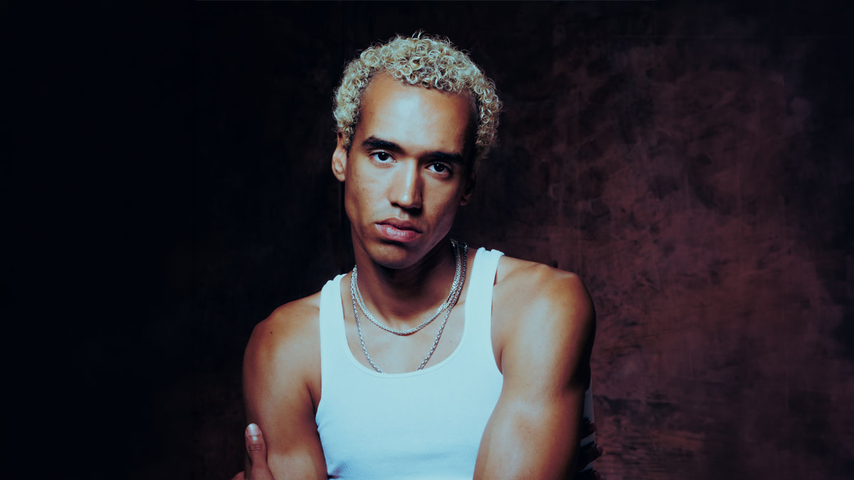 Portrait de WizTheMC, aux cheveux courts, blonds et bouclés, vêtu d’un débardeur blanc et de chaînes argentées, sur fond sombre.