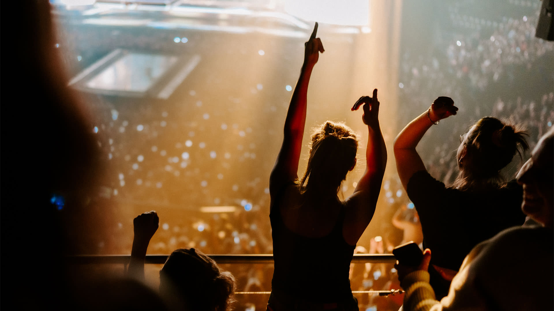 Publiek geniet van een concert in Vorst Nationaal, met fans die dansen en hun handen in de lucht steken terwijl de zaal oplicht door spots en telefoonschermen.