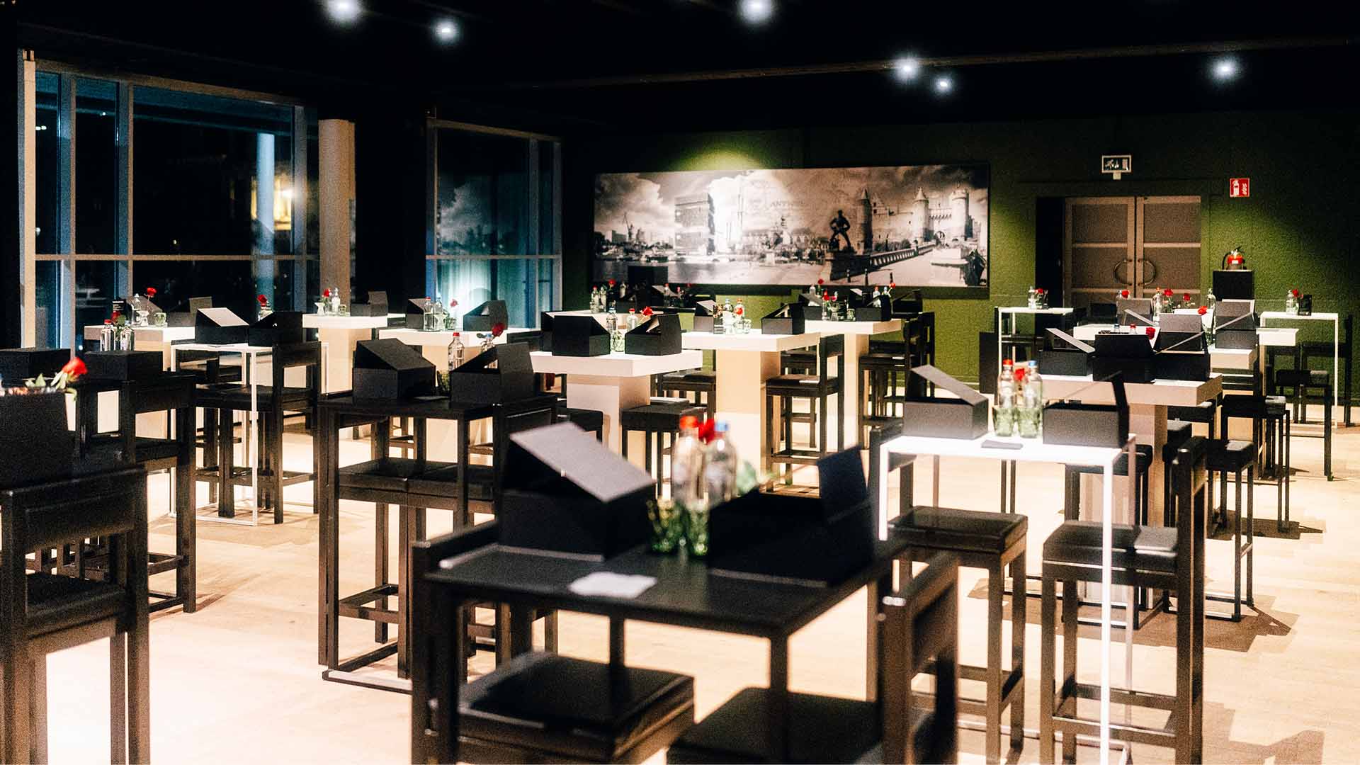 Een opgeruimde evenementenruimte met meerdere hoge tafels en stoelen, gedecoreerd met rode rozen en drankjes.