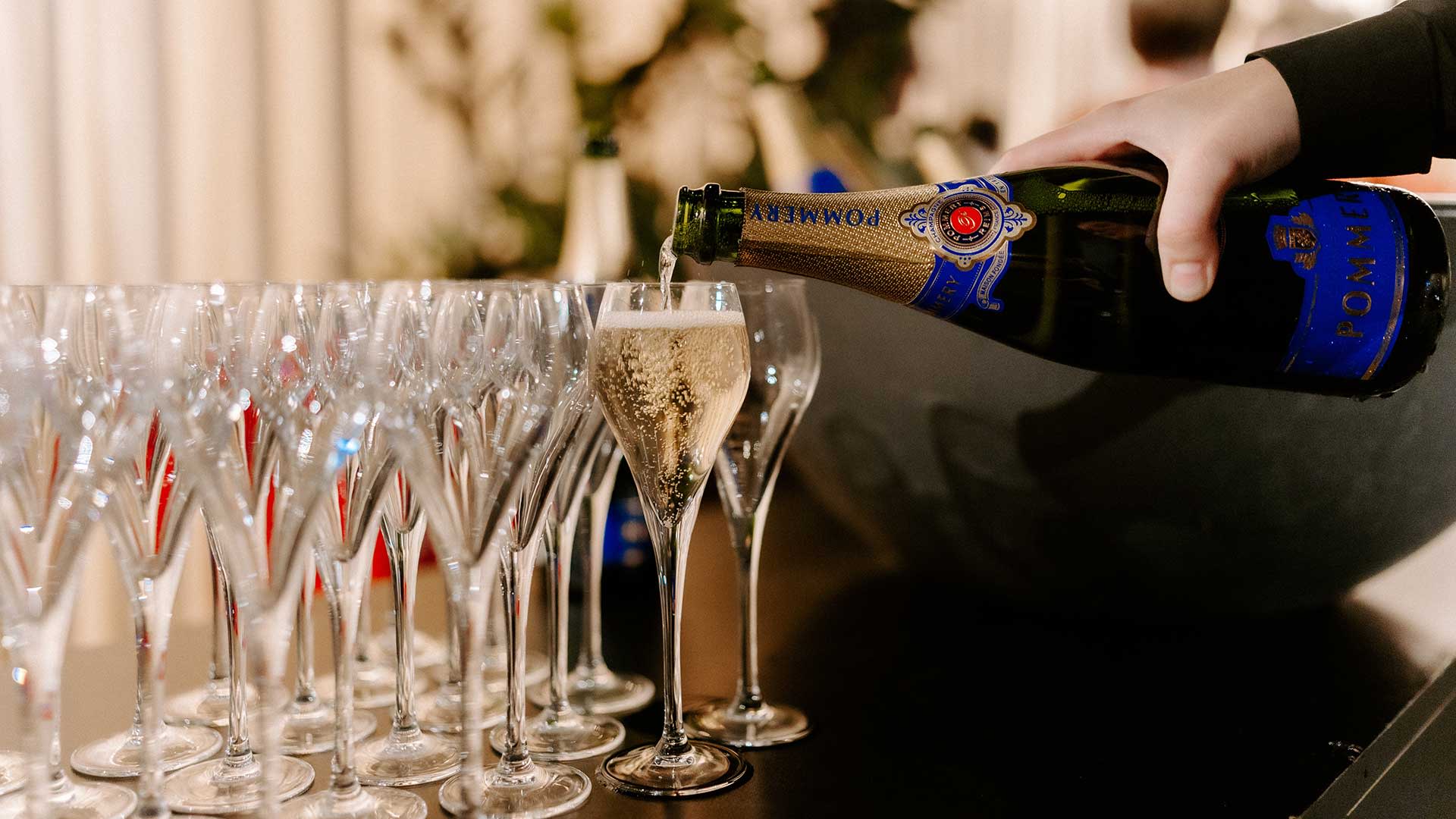 Plusieurs flûtes de champagne alignées sur une table pendant que le champagne est versé, avec des boissons pétillantes et un éclairage d’ambiance doux dans un espace lounge premium.