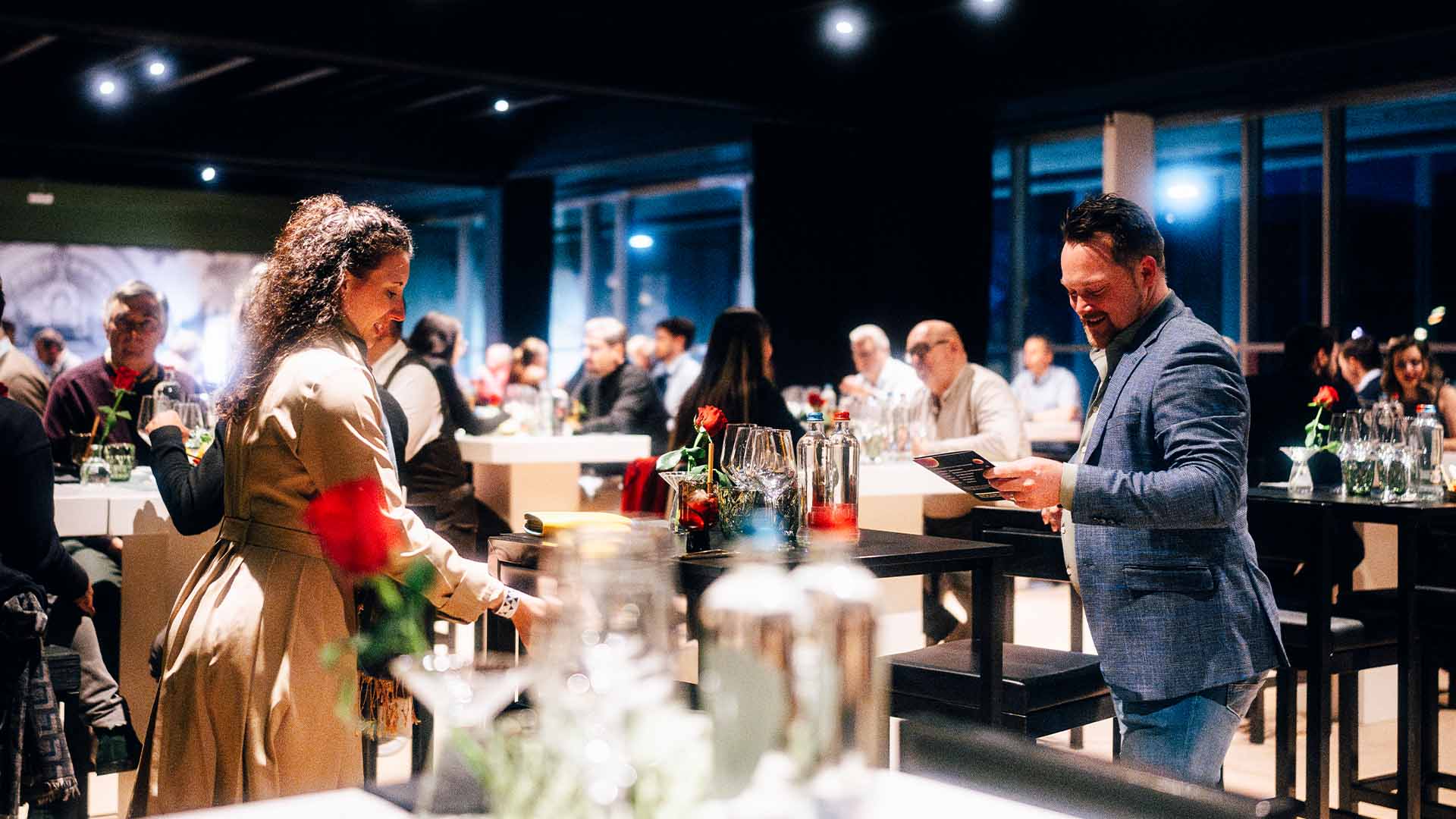 Un homme en veste bleue regarde un menu tandis qu'une femme en veste beige se tient près d'une table dans une pièce éclairée de manière atmosphérique avec d'autres invités.