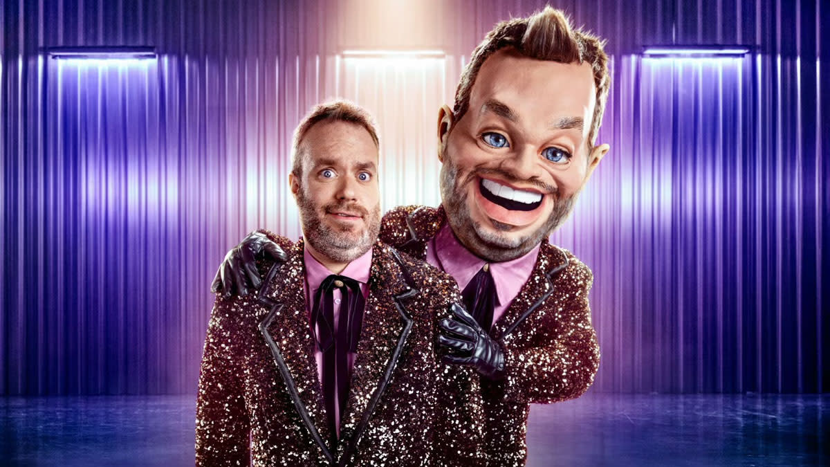 Presentator Jens Dendoncker naast zijn dubbelganger Grote Jens, een levensgrote pop met een lachend gezicht en glitterkostuum, in een kleurrijke studi setting.