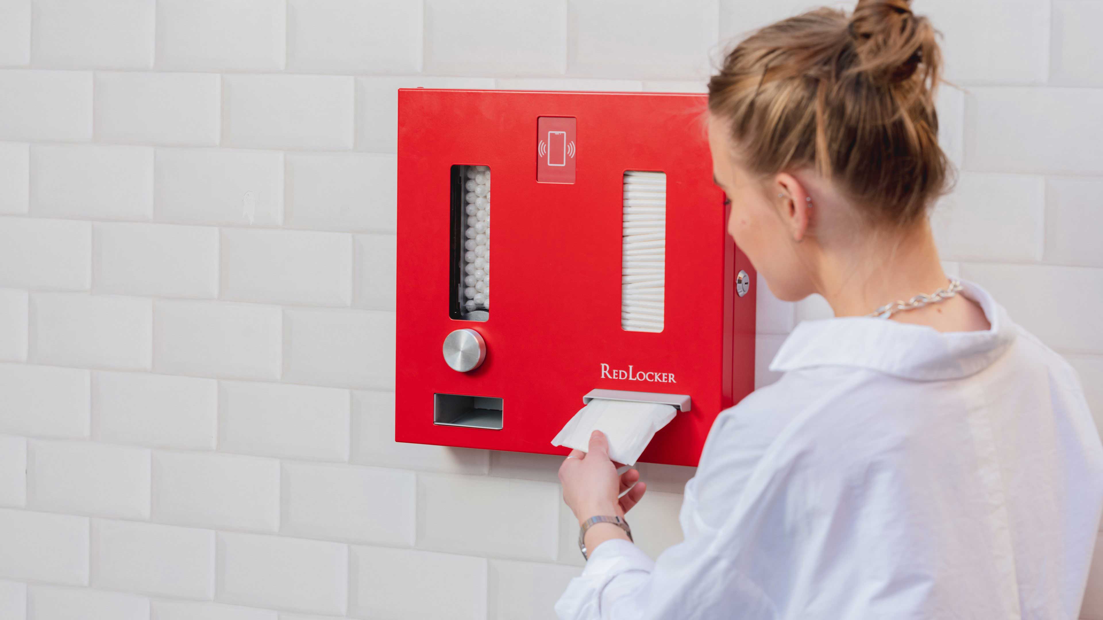 Een vrouw met blond haar, gekleed in een wit overhemd, haalt een maandverband uit een rode automaat met de tekst 'RedLocker' erop, die aan een witte tegelmuur hangt.