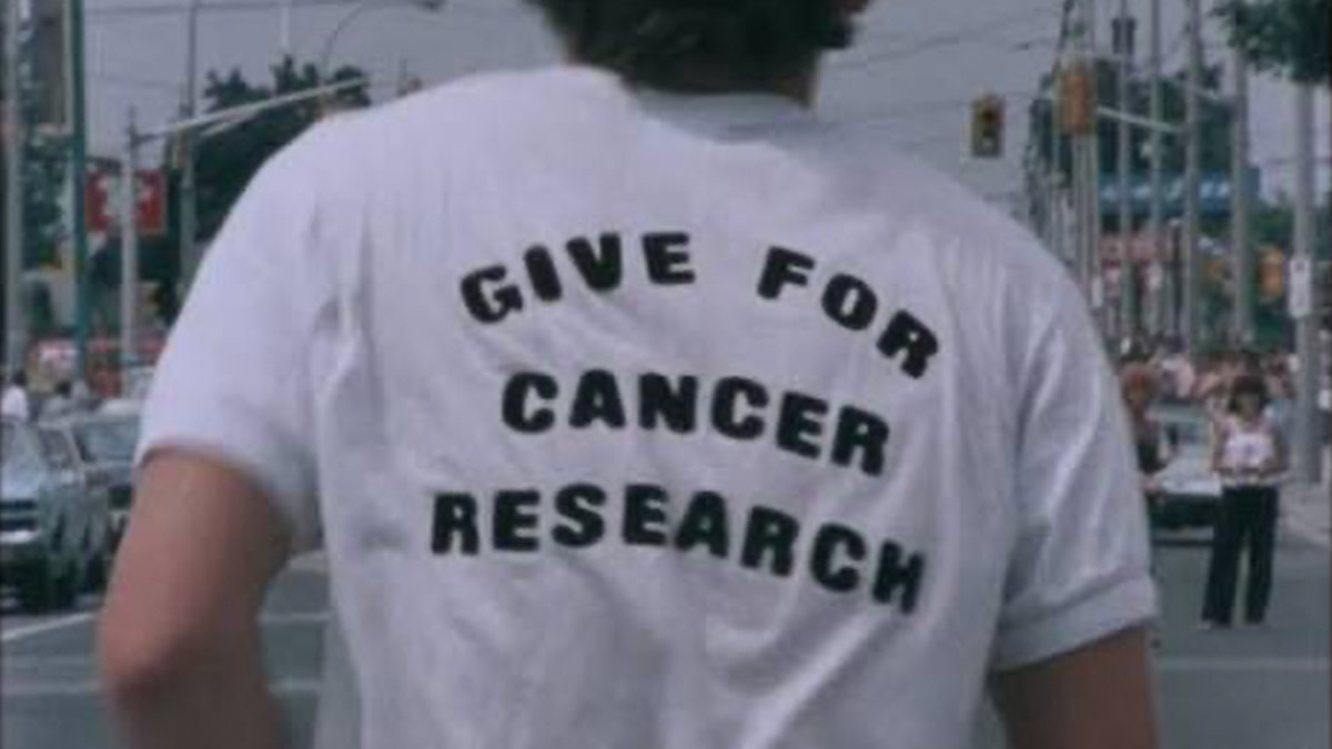 Terry Fox, filmed from behind on a street, wears a white T-shirt reading ‘Give for Cancer Research’.