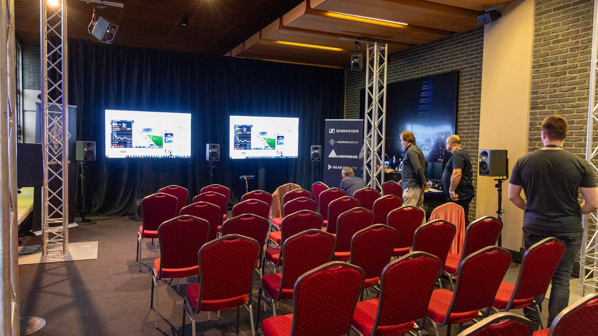 In de zaal staan verschillende rijen met rode stoelen opgesteld voor een presentatie. Vooraan in de zaal hangen twee schermen waarop de presentatie te zien is.