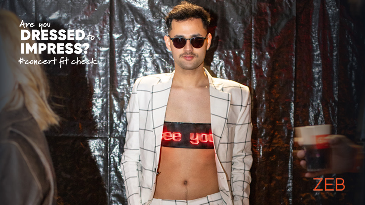 Een concertbezoeker poseert in een opvallende outfit met een wit kostuum met zwarte ruitjes en een lichtgevende top. Hij draagt zonnebril en kijkt zelfverzekerd in de lens.