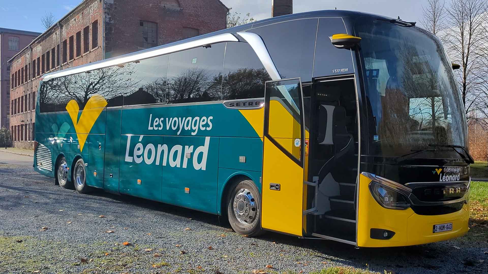 Autocar vert et jaune portant l’inscription « Les voyages Léonard », stationné sur un terrain ouvert près d’un ancien bâtiment en briques. La porte est ouverte et des arbres sans feuilles se trouvent à l’arrière-plan.