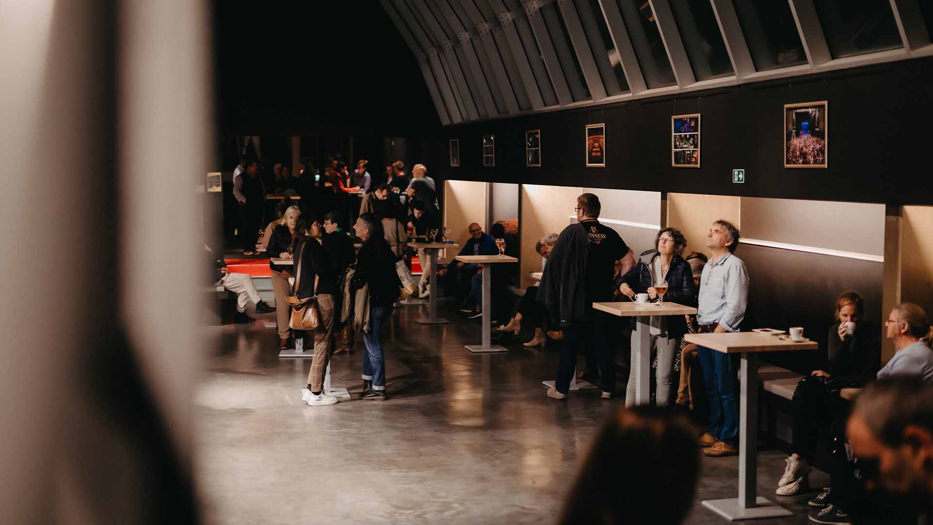 In de Koepelbar drinken verschillende mensen iets. Sommige staan aan hoge tafeltjes, andere zitten op een bank tegen de muur. Een deel van de koepel is te zien.