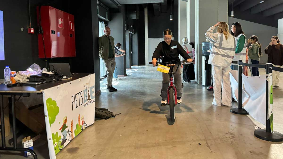 Een tiener fietst op een rode fiets door een hal. Op de achtergrond zijn andere mensen en een spandoek van FIETSPALEIS te zien.