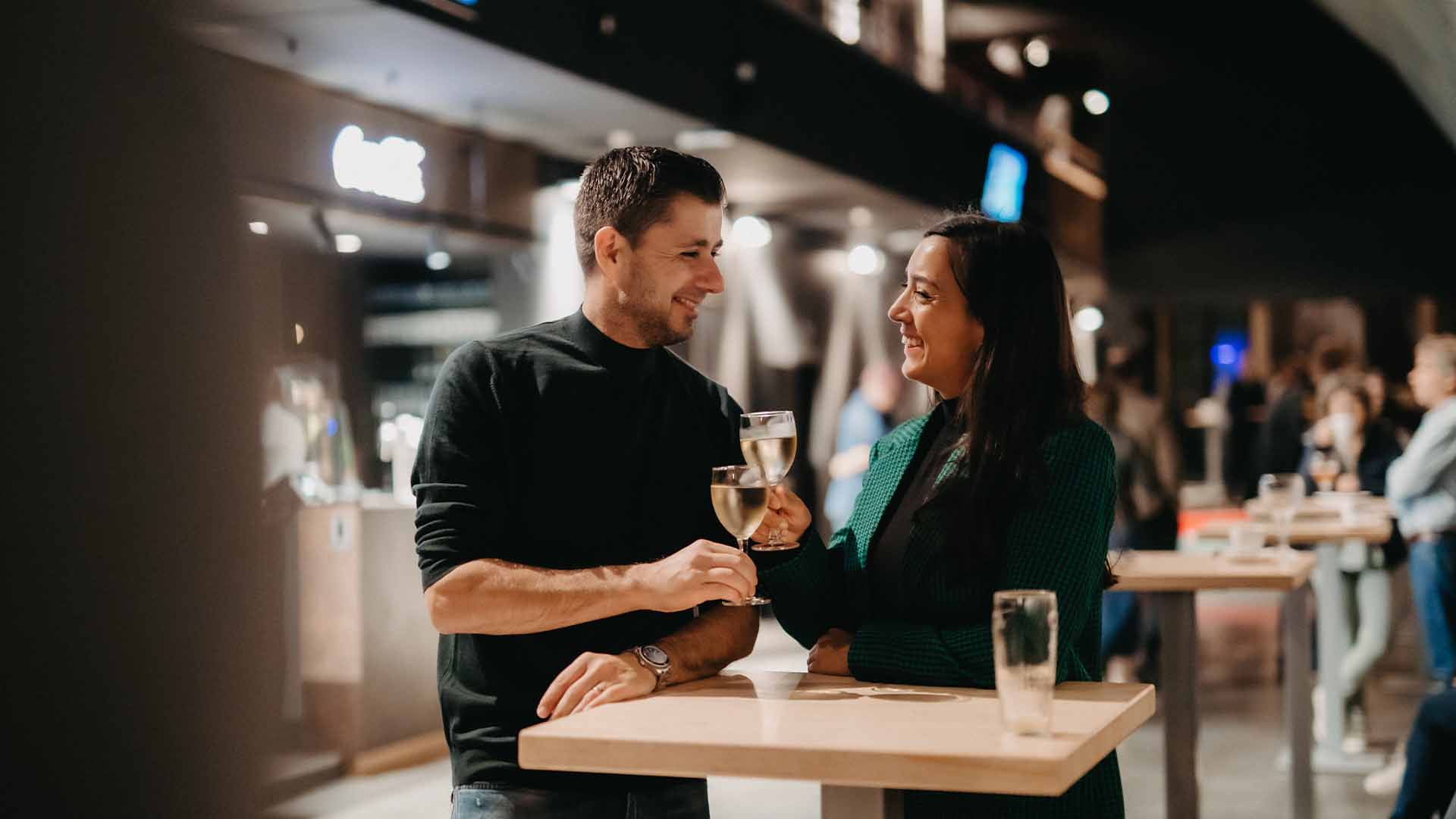 Een man en een vrouw kijken elkaar lachend aan en klinken met een glas witte wijn.