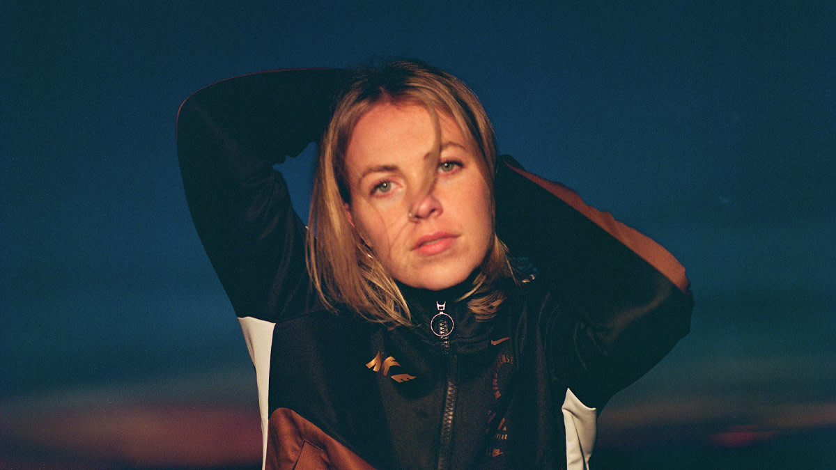 De Rotterdamse zangeres en songwriter philine kijkt in de camera terwijl ze haar handen in haar nek legt. Ze draagt een donker jasje en staat buiten bij schemerlicht, wat de foto een warme en dromerige sfeer geeft.