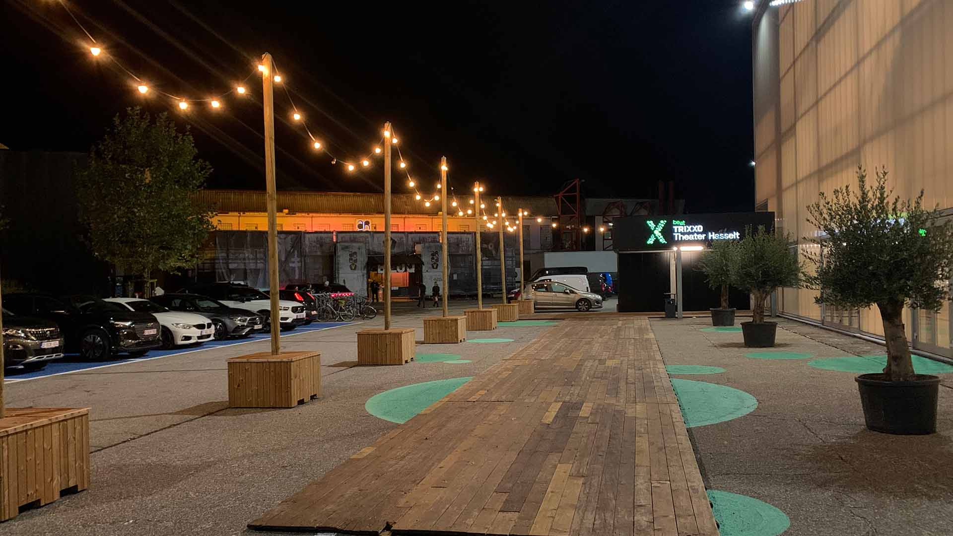 De verlichte buitenzone van Trixxo Theater Hasselt bij avond, met houten wandelpad, sfeerverlichting, planten in potten en geparkeerde auto's langs de zijkant.